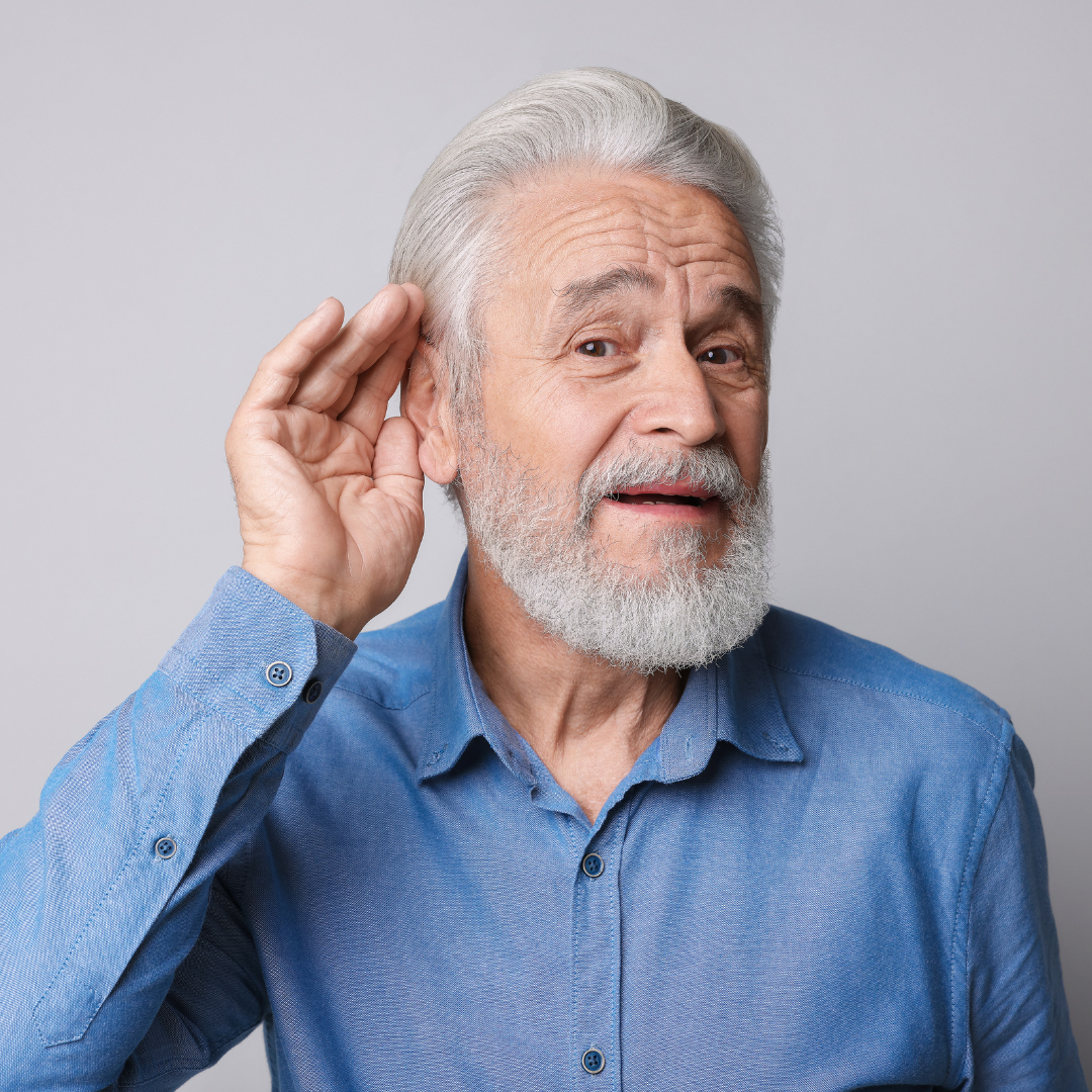 Older man with gray hair and beard wearing a blue shirt, holding one hand to his ear and tilting his head as if trying to hear better, against a plain light background.